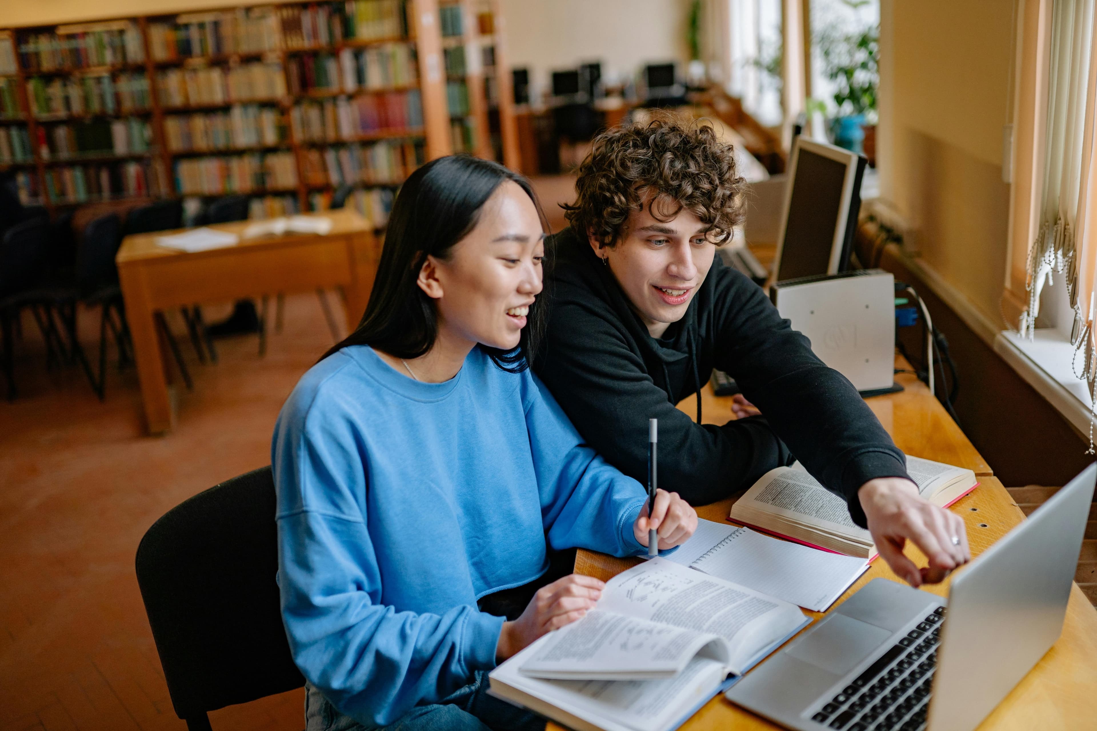 Dos estudiantes colaborando con libros y portátil en biblioteca